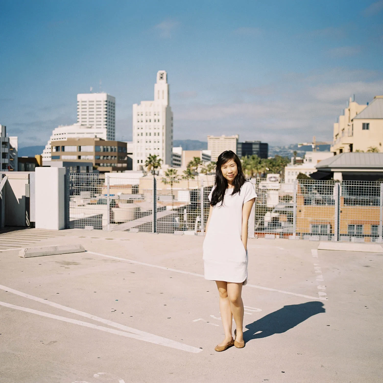 Woman in a pale dress standing on a rooftop with city towers behind her.