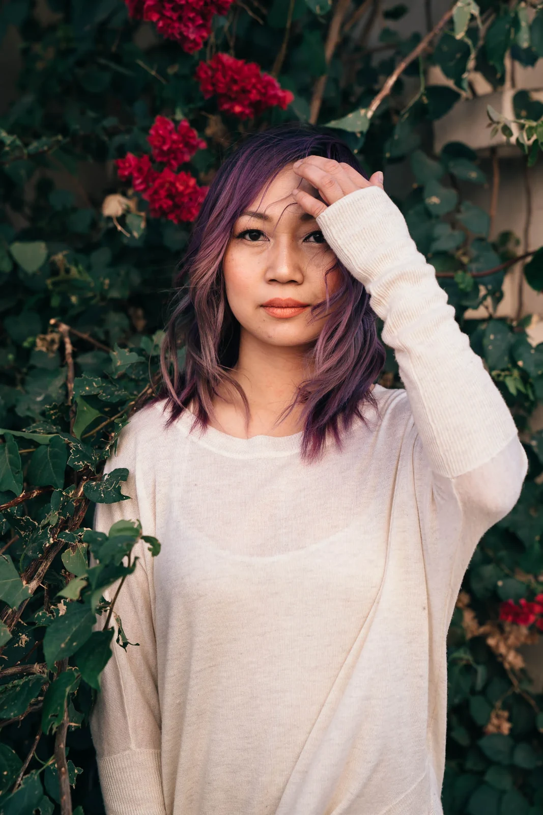 Woman with purple hair and a cream sweater standing before ivy and flowers.