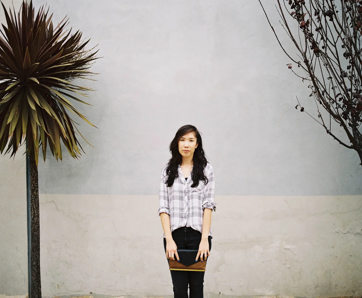 Woman in a plaid shirt standing against a pale wall between plants.