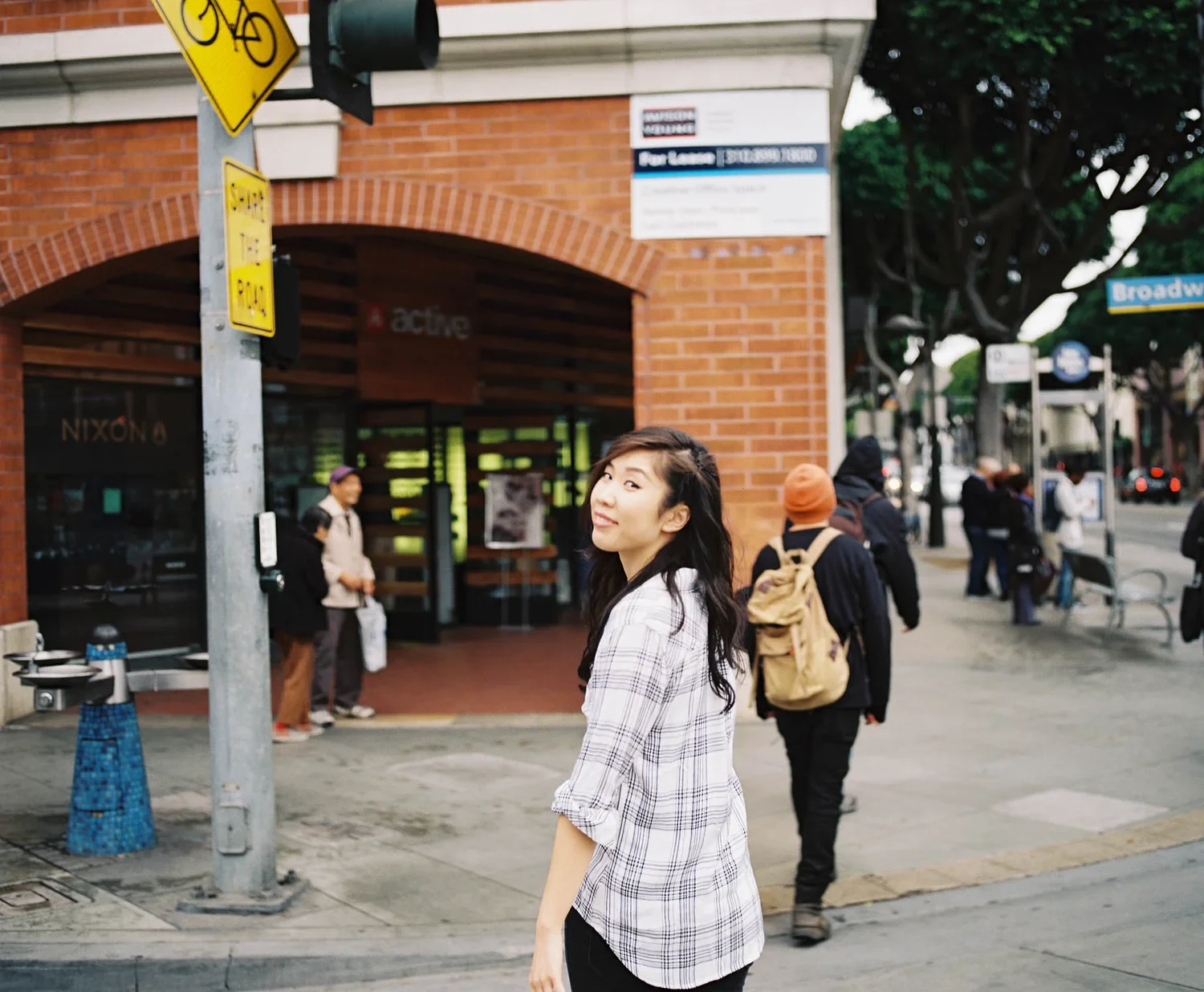 Woman in a plaid shirt looking upward on a city sidewalk.