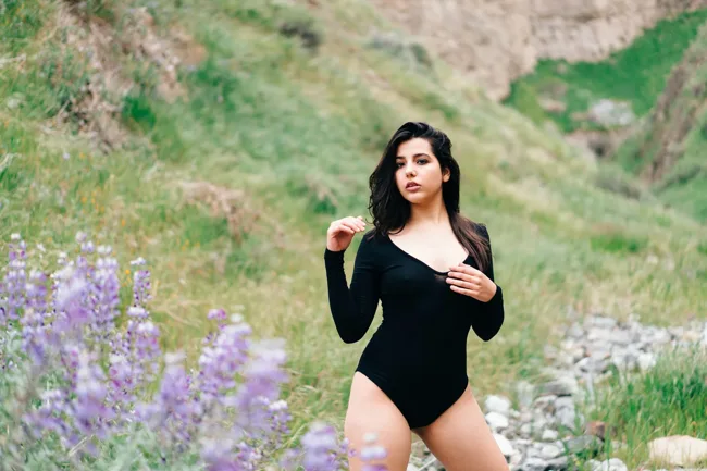 Model in black standing among purple wildflowers on a grassy hillside.