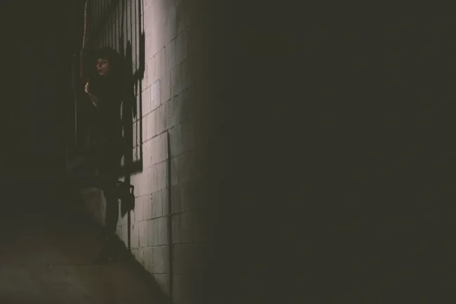 Dim portrait of a figure beside a narrow strip of light on a wall.