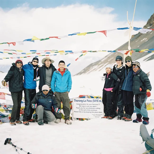 Group of trekkers posing beneath prayer flags on a snowy mountain pass.