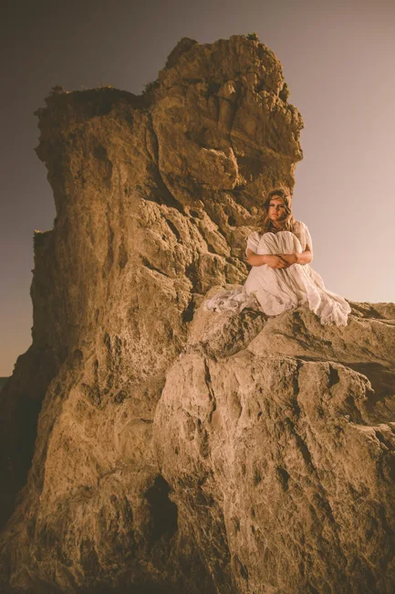 Model in white posing against a tall, sunlit rock formation.