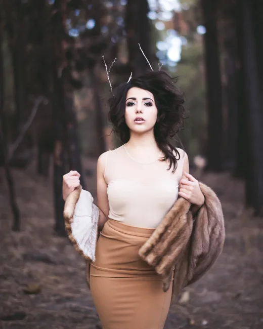 Model in a tan skirt and light top standing among trees.