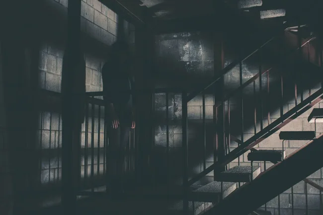 Model standing by industrial stairs amid a grid of window shadows.