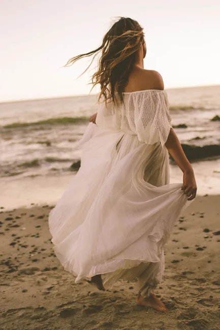 Model in a flowing white dress walking along a beach.