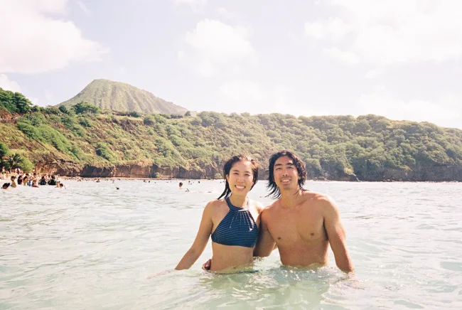 Two people standing together in ocean water with a green crater ridge behind them.