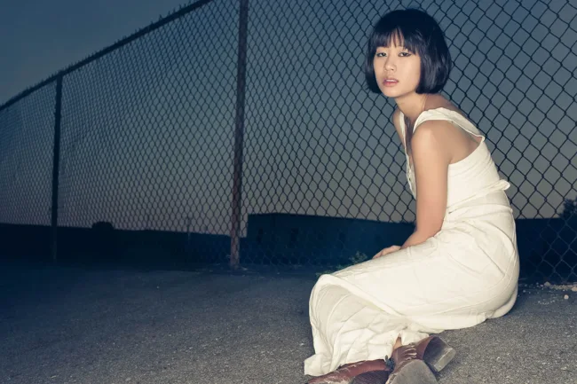 Model in a cream dress sitting beside a chain-link fence at dusk.