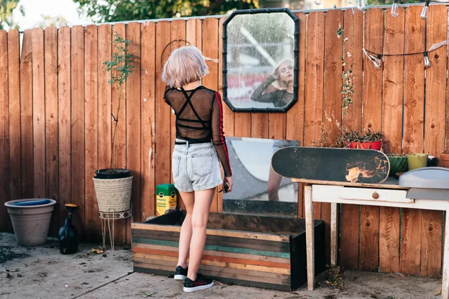 Model with pale hair standing beside a wooden fence and outdoor table.