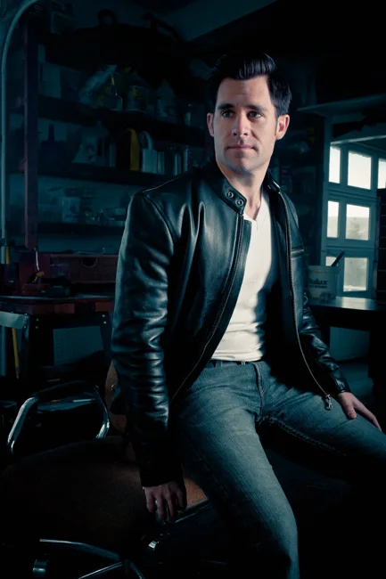 Man in a leather jacket seated in a dim workshop.