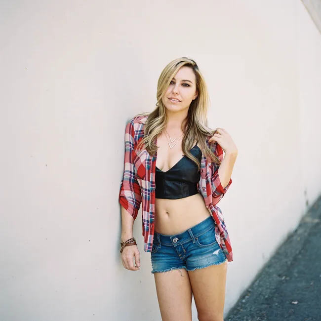 Woman in a plaid shirt and denim shorts smiling against a pale wall.