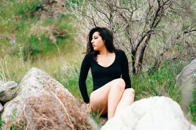Model in black seated beside large rocks and a small tree.