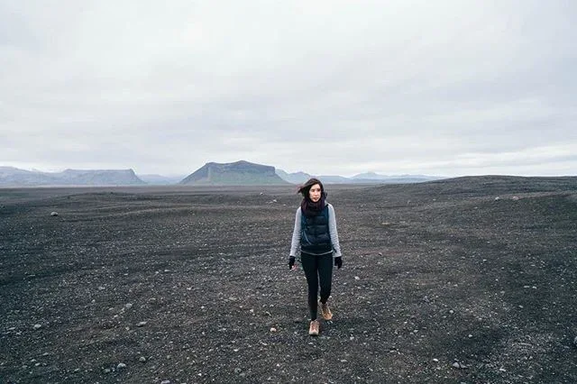 Woman standing on a wide black-sand landscape beneath an overcast sky.