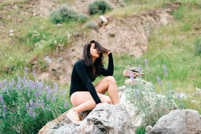 Model in a black bodysuit sitting among rocks and purple wildflowers.