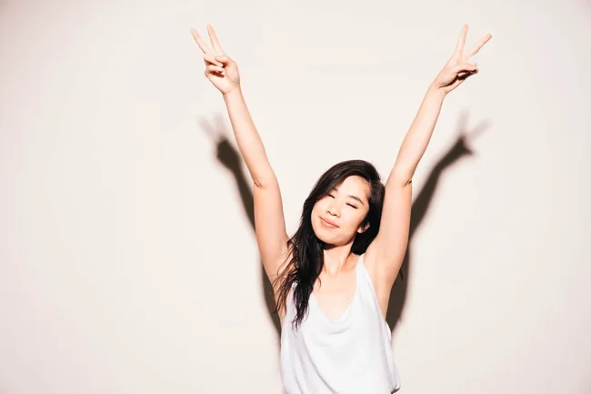 Model smiling with both arms raised against a pale wall.