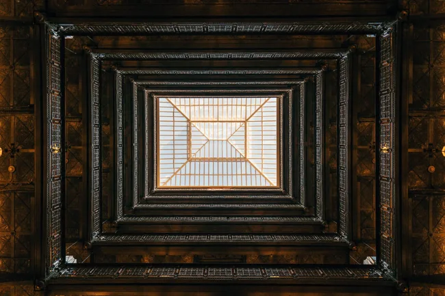 View upward through a square stairwell toward a bright skylight.
