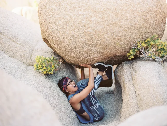 Person crouching beneath a huge boulder in a narrow rock opening.