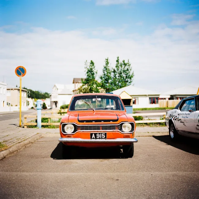 Orange vintage car parked beside houses in bright daylight.