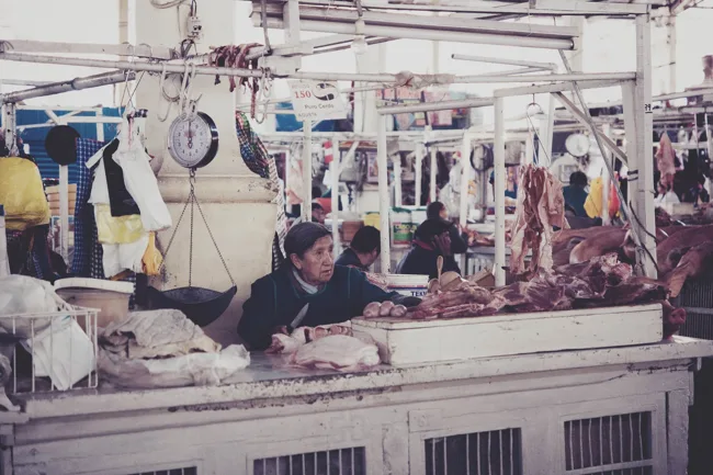 Vendor behind a counter beneath hanging cuts of meat in a market.