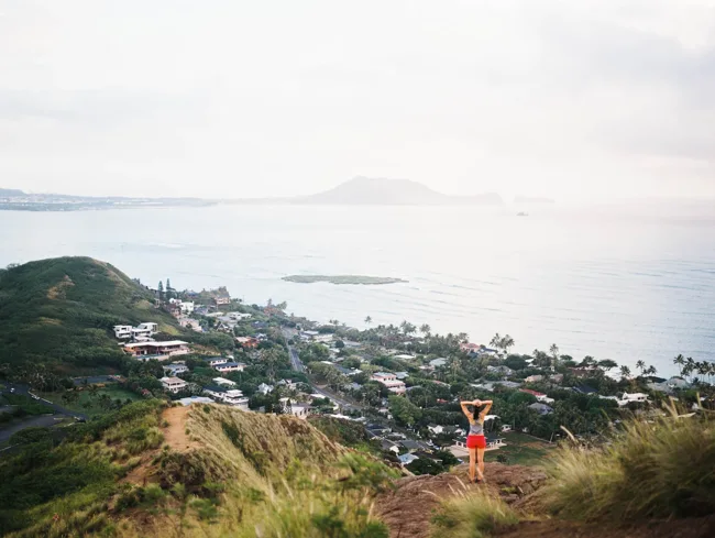 Hiker on a ridge overlooking a coastal town and island-dotted ocean.