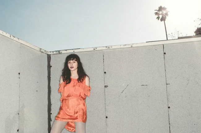 Woman in an orange dress standing beside a tall pale fence.