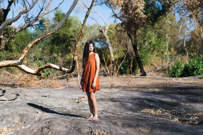 Woman in an orange dress and hat beneath spreading trees.