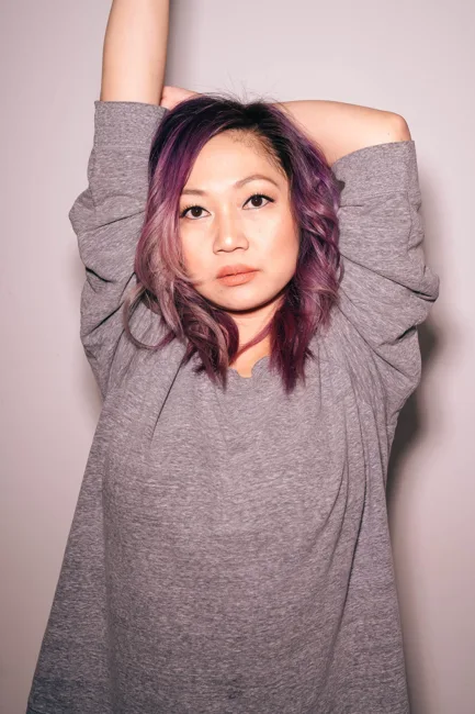 Model with purple hair raising her arms in a gray sweater.