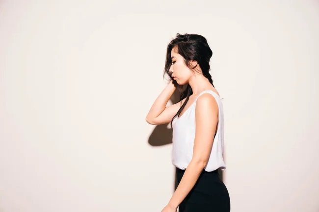 Model in a white tank top facing sideways against a pale wall.
