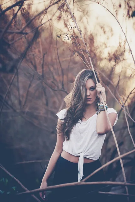 Model in a tied white shirt among branches in soft light.