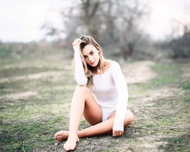 Model in a white bodysuit sitting on grass among bare trees.