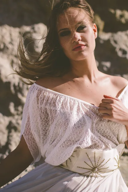 Model in a white off-shoulder dress beside coastal rocks.