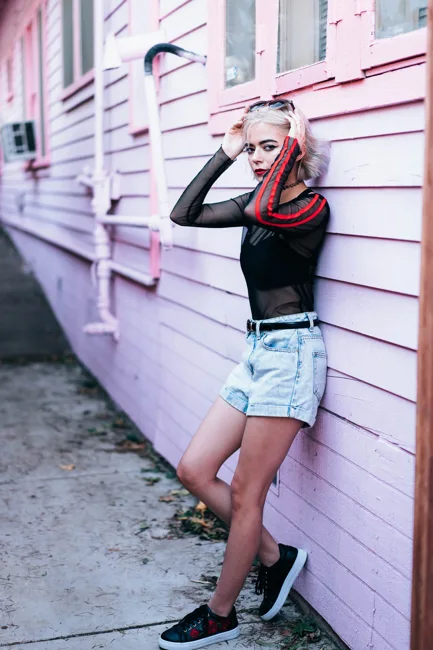 Model in denim shorts and a black top leaning against pink siding.
