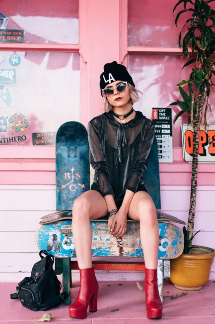 Model in sunglasses sitting with a skateboard in a pink storefront.