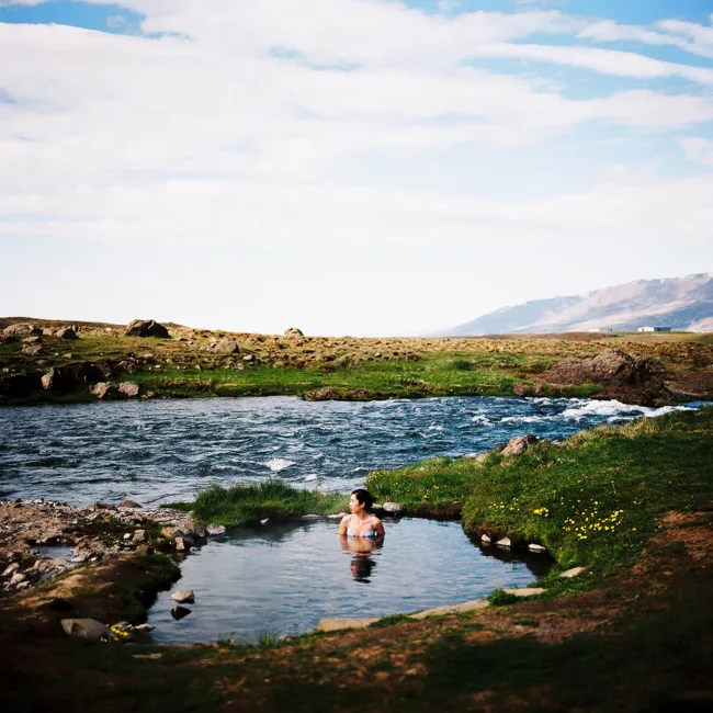 Person sitting in a small pool beside a river in an open landscape.