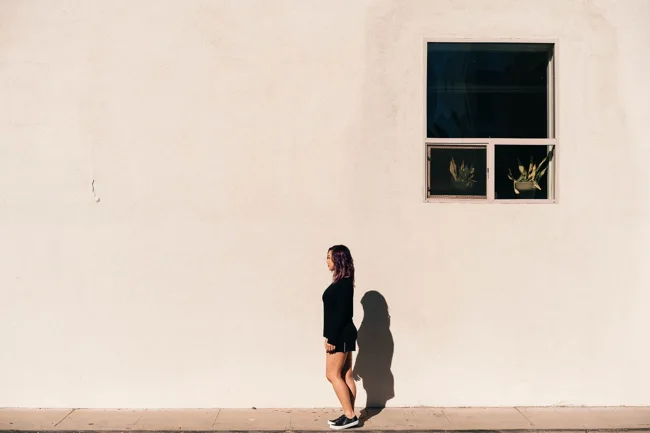 Woman walking past a pale wall beneath a small dark window.