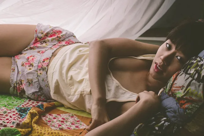 Model reclining on a floral-covered bed in soft light.