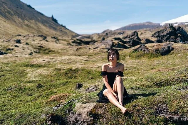 Woman in black seated among rocks in a grassy mountain valley.