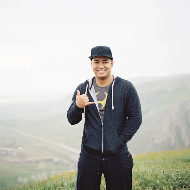 Man in a cap giving a thumbs-up in a misty mountain landscape.