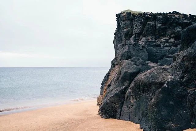 Dark coastal cliff above a pale beach and calm sea.