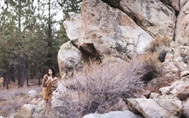 Model standing beside a large rock face at the edge of woodland.