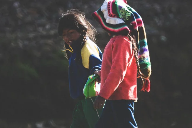Two people wearing colorful layers beside a shaded stone wall.