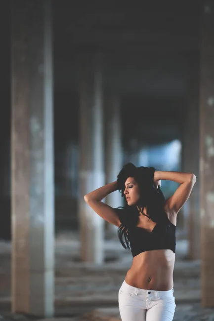 Woman posing with arms behind her head among dim concrete columns.